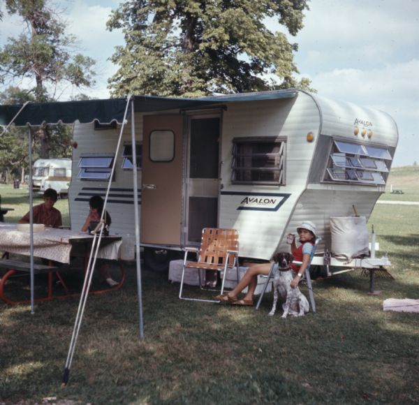 Three children are sitting at a picnic table, or in chairs next to an Avalon camper under an awning. A dog is sitting next to one of the girls.