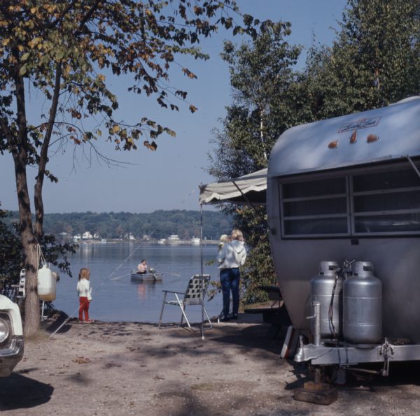 View towards a child, and a woman holding a baby at the shoreline of a lake. In the right foreground is a camper, and on the left is the front hood of a car. Out on the lake a man is rowing a metal rowboat on Long Lake in the Kettle Moraine State Forest.