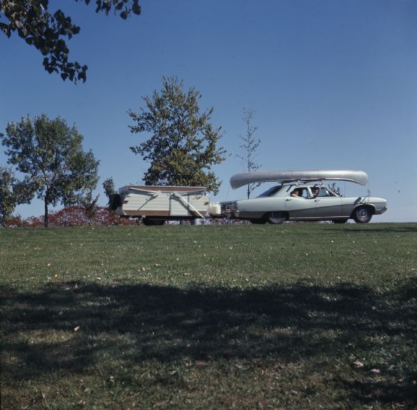 View across grass towards a group of people in a car pulling a camper, and a canoe strapped to the roof of the car, at a campground in Wildcat Mountain State Park.