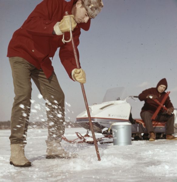 A man is standing on the ice of a lake, using an ice chisel to make a hole in the ice. Behind him a man is sitting on an Evinrude snowmobile prepping the tip up.