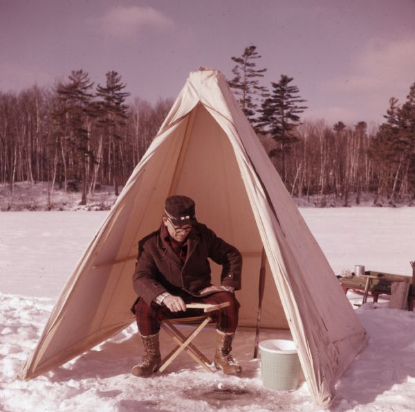 A man is sitting on a stool in the open doorway of a small tent on the ice on Lake Owen. He is holding a wooden drop line over a hole in the ice. Behind the tent is a sleigh, and trees are along the shoreline.