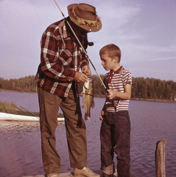 A man is showing a young boy how to unhook a bass from a fishing line while standing on a pier. The boy is holding the fishing pole. The man is wearing a fishing hat and is smoking a pipe.