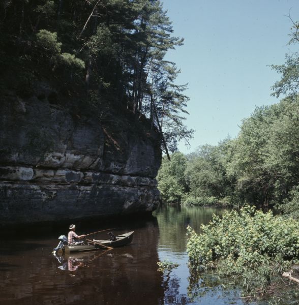 A man is sitting in a boat holding a fishing pole on the Wisconsin River near Tower Hill State Park. A black dog is sitting towards the front of the boat.