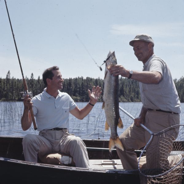 Two men in a canoe on a lake, with one of the men holding a muskellunge (musky) hooked on a fishing line, and a fishing net. The other man is holding the fishing pole with the musky.