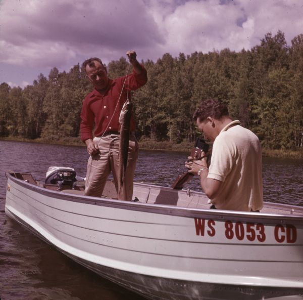 A man is standing in a boat on Pine Lake, holding up a muskellunge he has caught. Another man is sitting in the boat holding a camera to take a photograph of the fish.