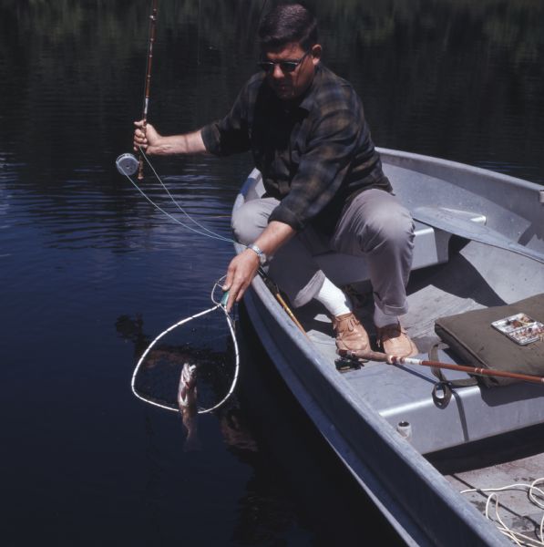 Close-up of a man leaning over the side of a boat and using a small net to scoop up a trout he has caught on the line of his fly rod.