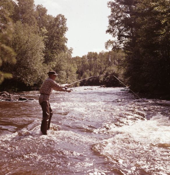 View across water towards an elderly man standing calf-deep in the Bois Brule River holding fly rod.