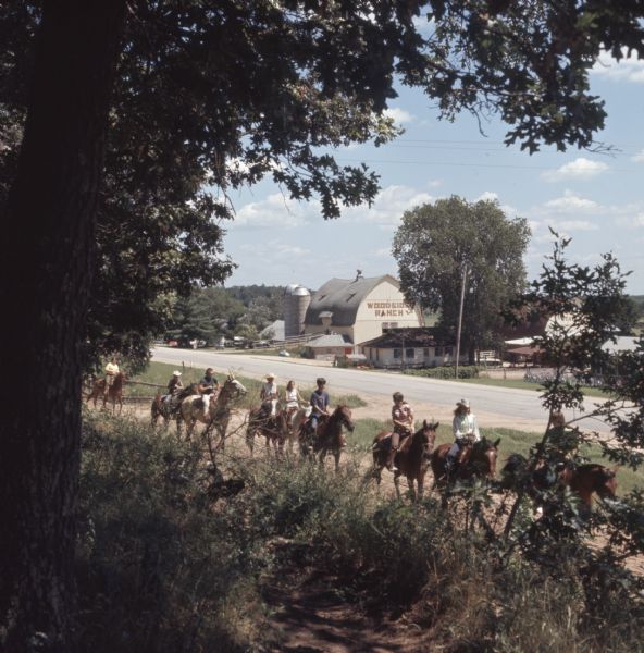 View from shade of trees towards a line of teenagers and adults riding horses along a dirt path. Across the road in the background are the barns and stables of Woodside Ranch.