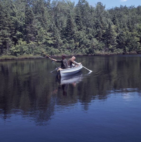 View across water towards two men fishing from a rowboat. Trees are along the opposite shoreline.