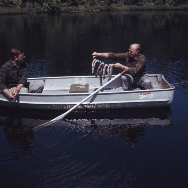 Slightly elevated view of two men sitting in a rowboat. One of the men is holding up a stringer of fish.