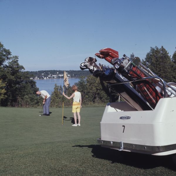 A man and woman are on the putting green of a golf course, with a golf cart in the foreground on the right, and in the background a view of the bay. The woman is standing and holding the flag stick as the man is preparing to putt the golf ball.