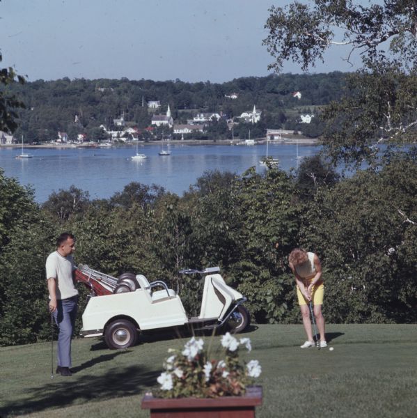 A woman getting ready to putt on the green at the Peninsula State Park Golf Course. A man stands on the left watching, and parked behind him is a golf cart. In the background, sailboats are moored in the bay, and there is a town on the far shoreline.