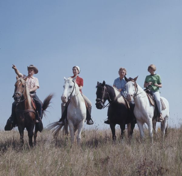 A group of four horseback riders in tall grass on the top of a hill in Kettle Moraine State Forest. The man on the left is pointing off into the distance.