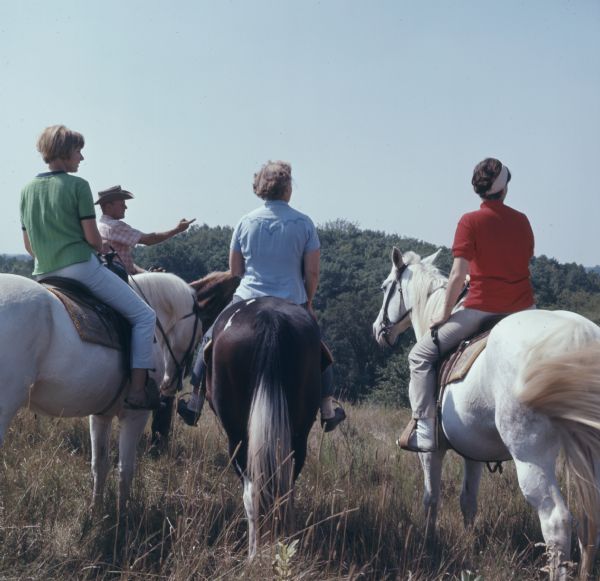 A group of four horseback riders, one man, and three women, are stopped at the top of a hill looking at the view at Kettle Moraine State Forest. The man on the left is pointing to the right.