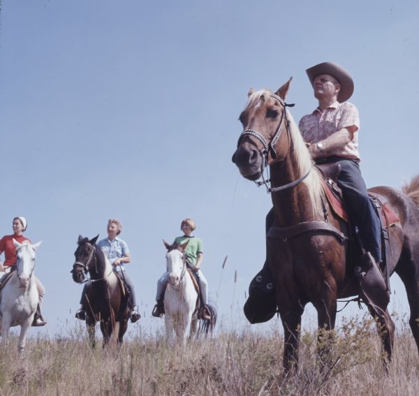 A man in the foreground on the right is sitting on horseback at the top of a hill. In the background on the left are three women on horseback.