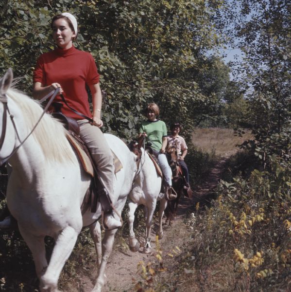 Two young women and a man are riding horses single file on a narrow dirt path in the Kettle Moraine State Forest.