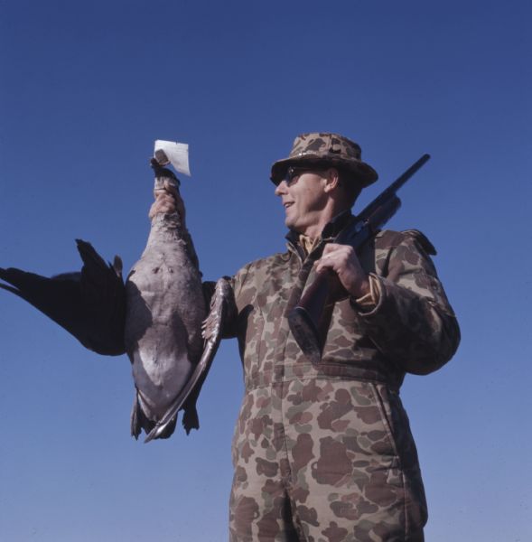 A hunter is holding his rifle over his left shoulder and is holding a goose in his right hand.