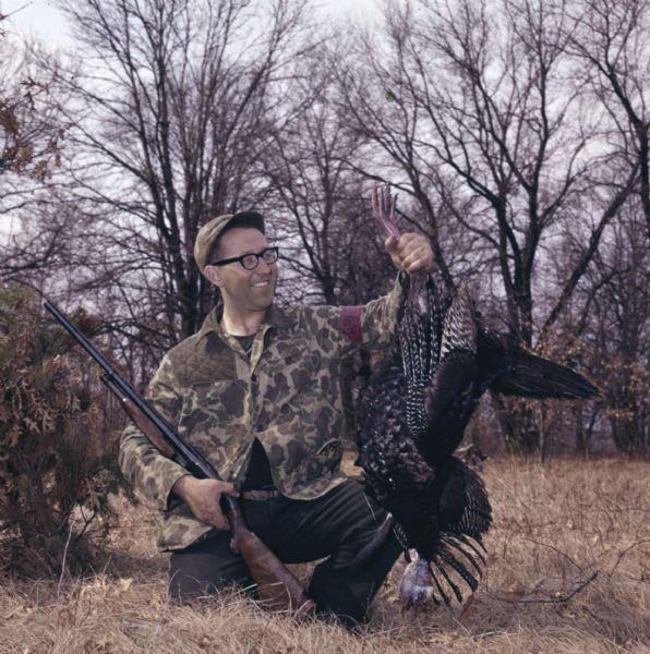 A man in hunting gear is kneeling on the ground holding his rifle in his right hand, and holding up a dead turkey in his left.
