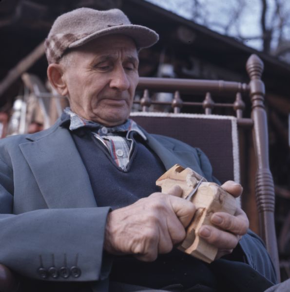 Close-up view of a man sitting in a chair and whittling an animal figure.