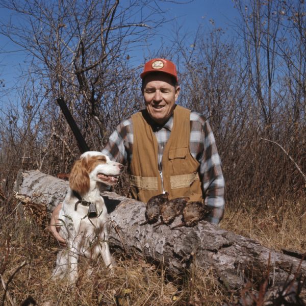 View of a man kneeling behind a log, with his arm around his hunting dog. Three dead woodcocks are lying on the log in front of the man.