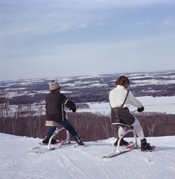 View of a boy and girl at the top of a snow-covered hill sitting on skibobs, ready to go down the hill.