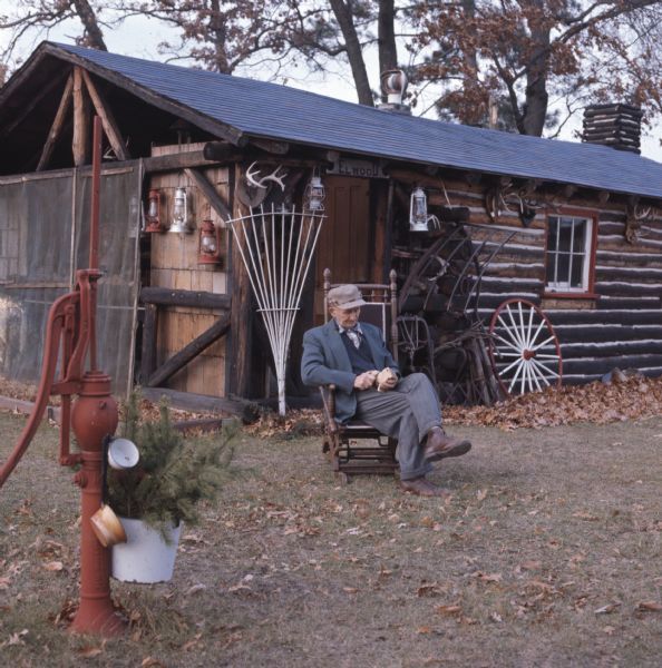 Man sitting on a rocking chair in a yard whittling in a rocking chair in front of a log cabin that has a sign reading: "Elwood." Lanterns and antlers are hanging on the side of the cabin, and in the foreground is a bucket of pine branches hanging on a red water pump.
