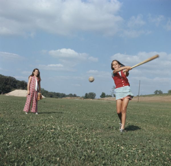 Two young girls are playing softball in an open grass field. One girl is swinging a bat at a ball flying in midair. The other girl is standing behind her on the left wearing a catchers mitt.