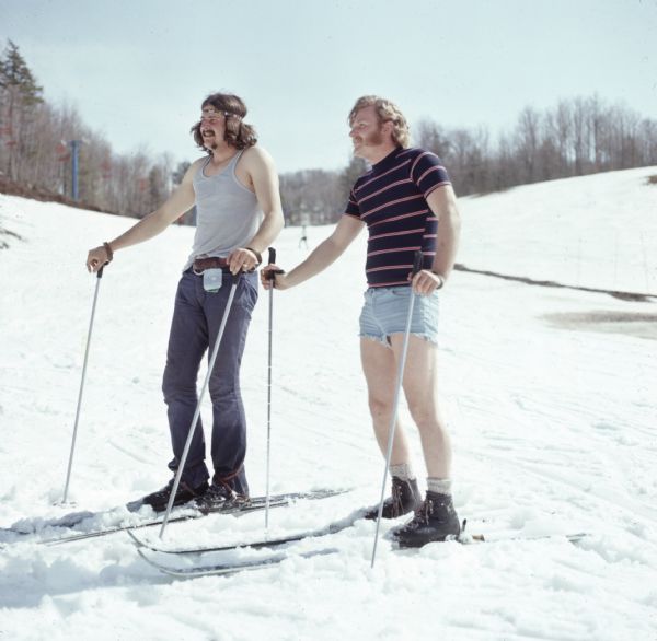 Two men wearing skis are standing on snow. One is wearing a sleeveless shirt and jeans, and has shoulder-length hair held back with a headband. The other man, who has mutton chops, is wearing a t-shirt and shorts.