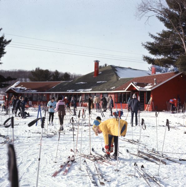 People outdoors in front of the ski lodge at Mt. Hardscrabble. Ski poles are standing upright near skis left in the snow in the foreground.