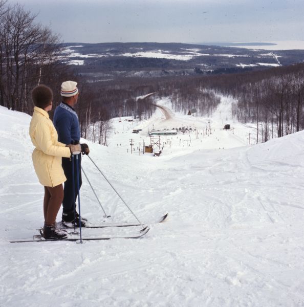 A man and a woman are standing on their skis looking down the ski hill looking down at the landscape below.