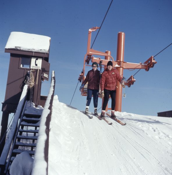 View looking up towards a man and a woman just getting off the ski lift at Mt. Telemark Resort. An emergency telephone booth sign is on the booth next to the hill on the left.