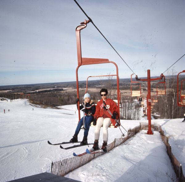 Slightly elevated view of two young people riding the chair lift at a ski hill. 