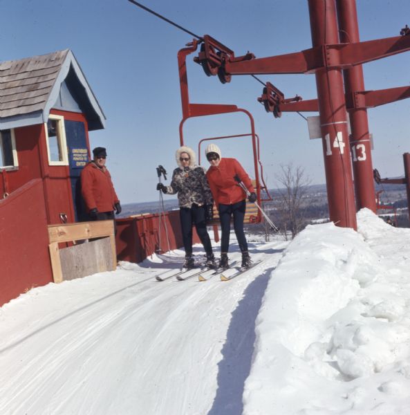 Two women are getting off the chair lift at the top of the hill at Mt. Telemark Resort. A man is standing next to a small red booth on the left.