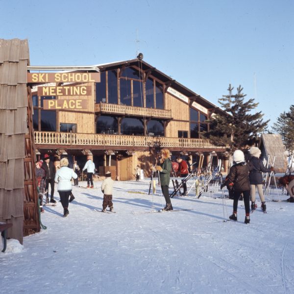 Men, women, and children on the snow outside a lodge. Skis are stacked on racks. A sign on the left reads: "Ski School Meeting Place."