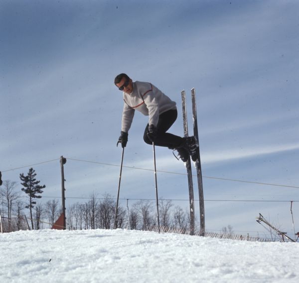 A man is holding his body off the snow-covered ground, supporting his weight entirely on the tips of his skis and ski poles.