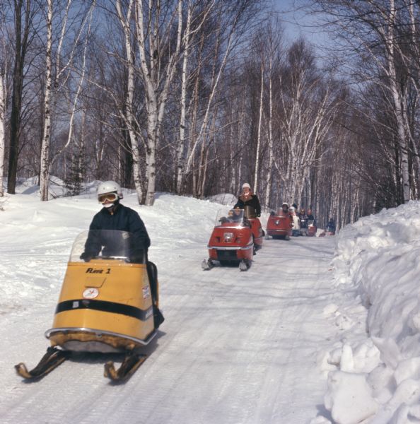 View towards a group of people riding snowmobiles through a path in the woods.