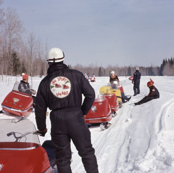 A group of snowmobilers are resting in the snow or on their snowmobiles. One person in the foreground is wearing a snowsuit with a large insignia on the back that reads: "Sno Sport Safaris."