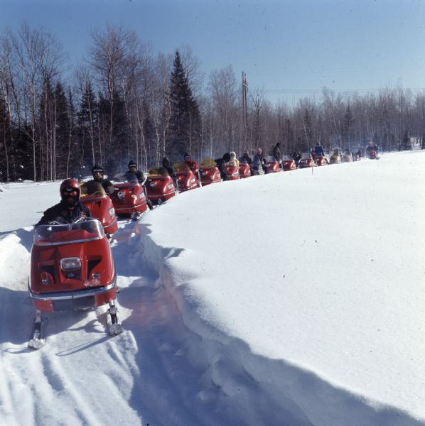 View looking towards a group of people driving snowmobiles down a hill  on a path through deep snow.
