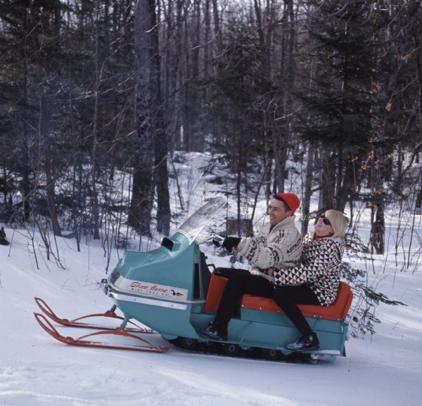 A man and woman are riding a teal and orange snowmobile through the woods.