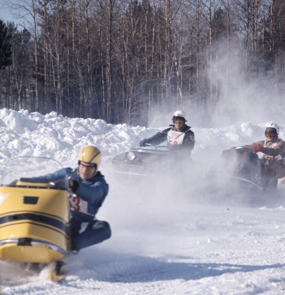Three men racing snowmobiles in the Winterama race. Each man is wearing a cloth bib with a race number on it.