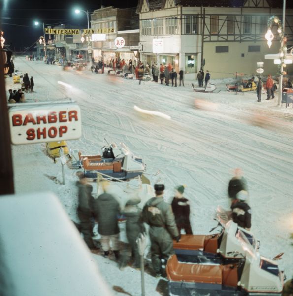 Elevated view of a crowd of people with snowmobiles standing on both sides of a snowy street downtown. There is a sign for a barbershop on the left, and a banner hanging over the street reads: "Winterama Jan 11-14."