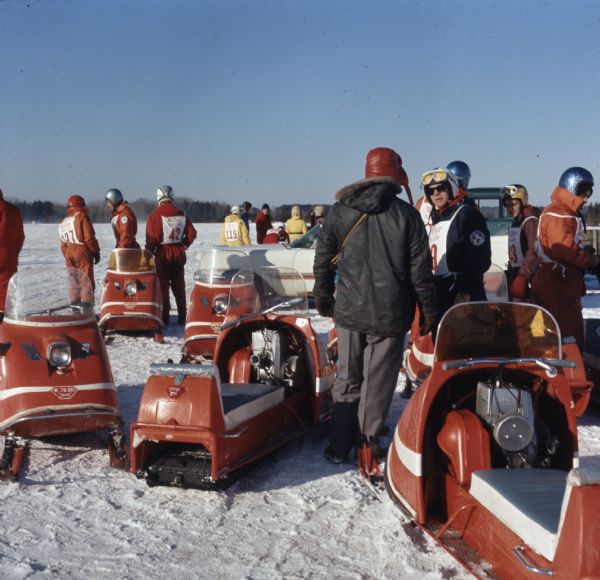 Men wearing helmets and race numbers are standing outside in the snow next to red snowmobiles.