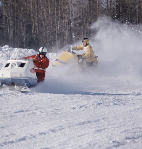 Two people wearing race numbers are riding snowmobiles around a turn, creating a cloud of snow behind them.