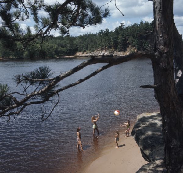 View from rock cliff looking down at four girls standing on a beach in a square, throwing a beach ball to each other. The older girls are standing in the water at the edge of the water of the Wisconsin River. The two younger girls are standing on the sandy beach.