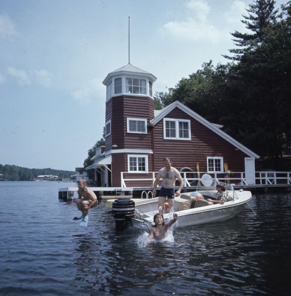 View across water towards two children jumping off the back of a motor boat into the water. The mother and father are watching from the motor boat. Behind them is a large red boathouse with a turret.