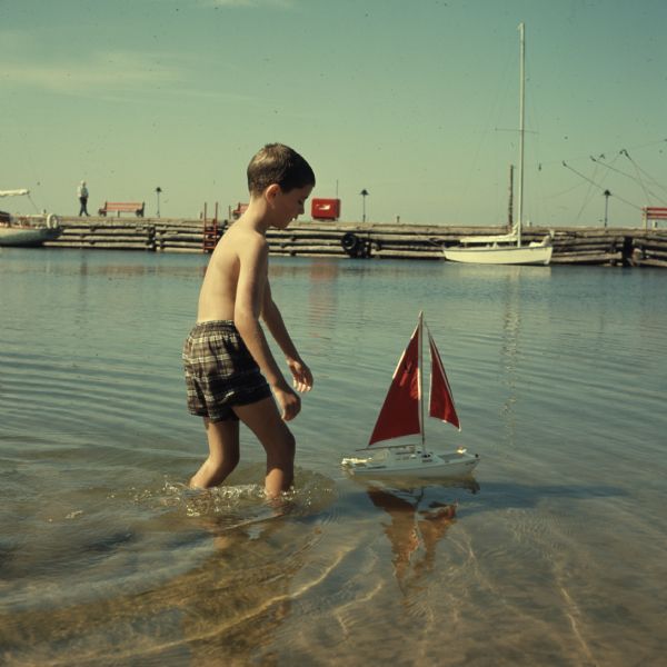 A boy is wading through the shallow water of Lake Michigan following a toy sailboat. In the background are sailboats tied to a dock at Gordon Lodge.