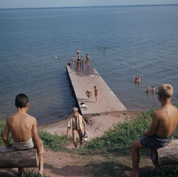View looking downhill between two boys in the foreground sitting on logs on the left and right. Another person is walking up the hill path. Below is a group of young people standing on a dock in Chequamegon Bay, some preparing to jump in the water, and others swimming or standing in the water.