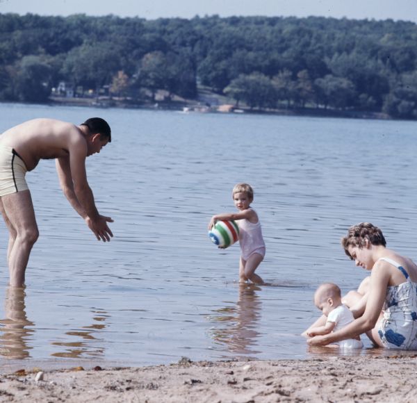 A woman and a baby are sitting in the shallow water on a sandy shoreline. A man and child are playing with a beach ball nearby further out in the water. The opposite shoreline is in the background.
