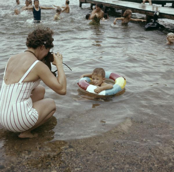 A mother is kneeling on the edge of Timms Lake, photographing her son who is floating in an inner tube. Other children are playing in the water in the background near a pier.