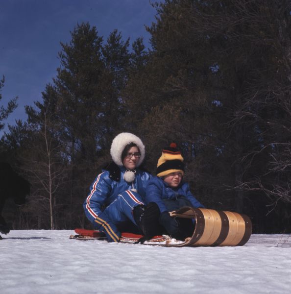 A young woman and a child are sitting on a toboggan. On the left is a black lab running towards them.